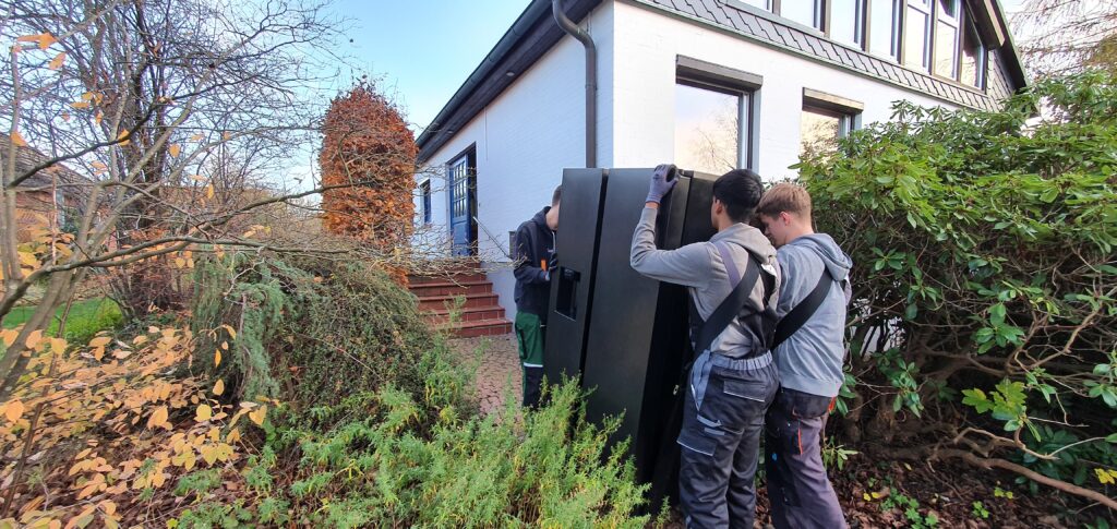 alt="Umzugshelfer transportieren einen Kühlschrank mit Tragegurten in ein Haus"